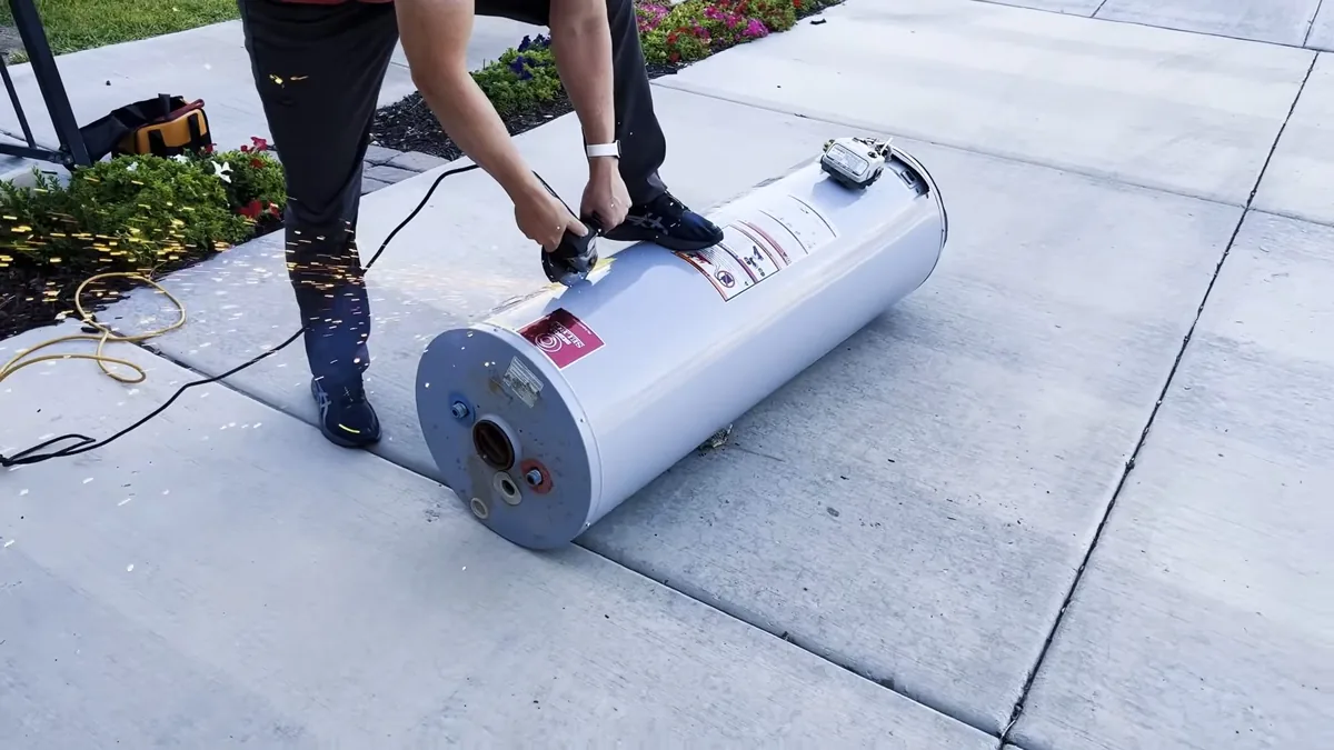 Person in dark clothing rolling a tank water heater across a concrete sidewalk beside flower beds